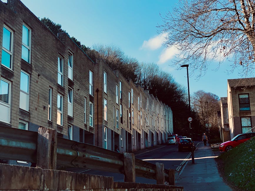 A residential area along King Georges Road featuring modern multi-story apartment buildings constructed with light-colored bricks and large windows. The scene includes a sloped sidewalk with a parked 