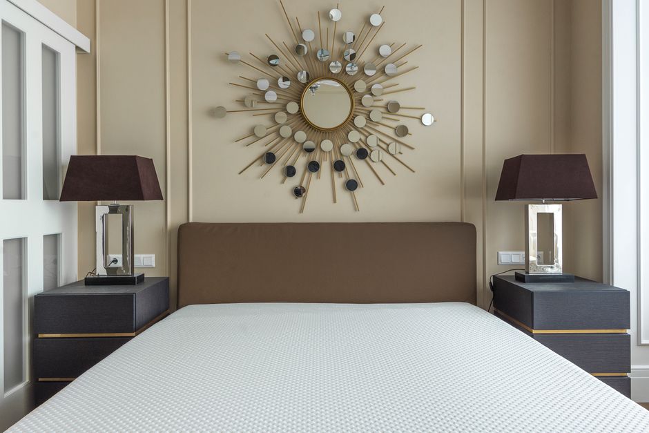 A neatly made white bed with a padded headboard in a modern, minimalistic bedroom featuring white walls and a closed beige wardrobe with silver handles in the background. The room is brightly lit by n